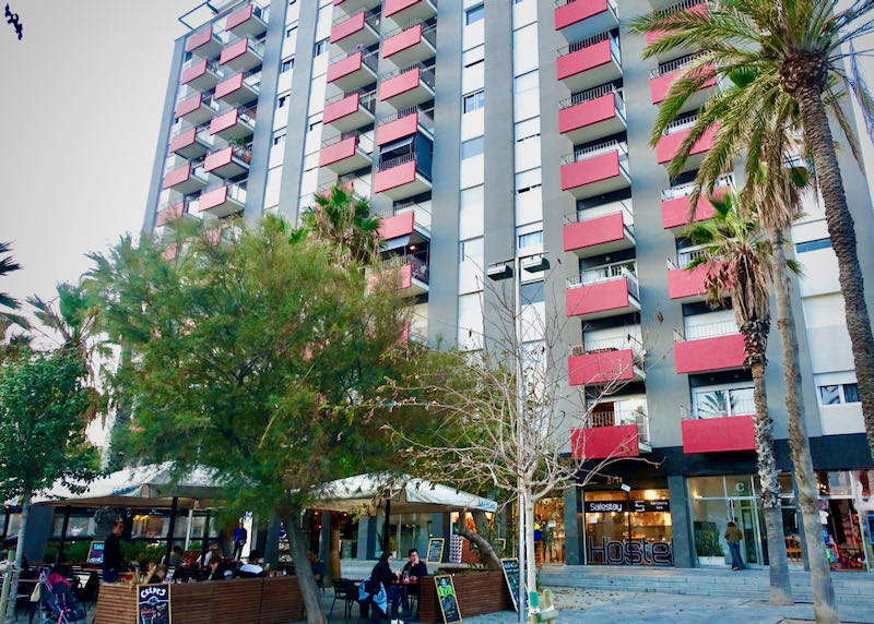 Hotel de gran altura con balcones rojos y una cafetería en frente