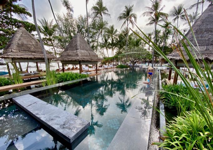 Dos mujeres descansando cerca de una piscina serpenteante bordeada de palmeras y cabañas de estilo tailandés