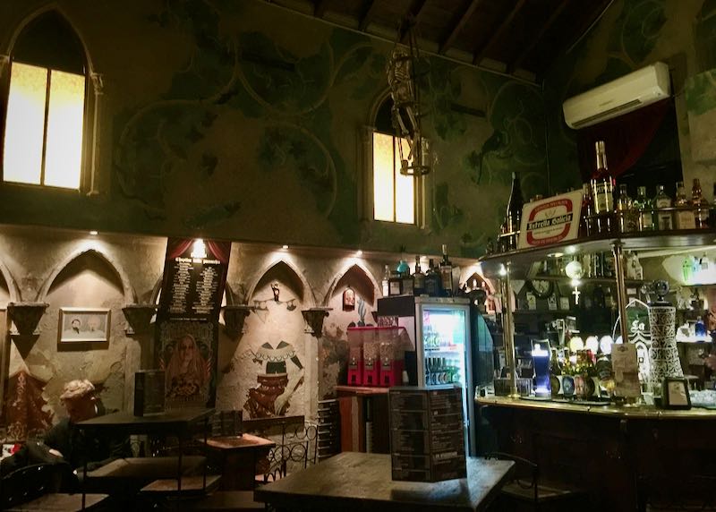 Interior con poca luz de un bar con ventanas góticas arqueadas y frescos en la pared