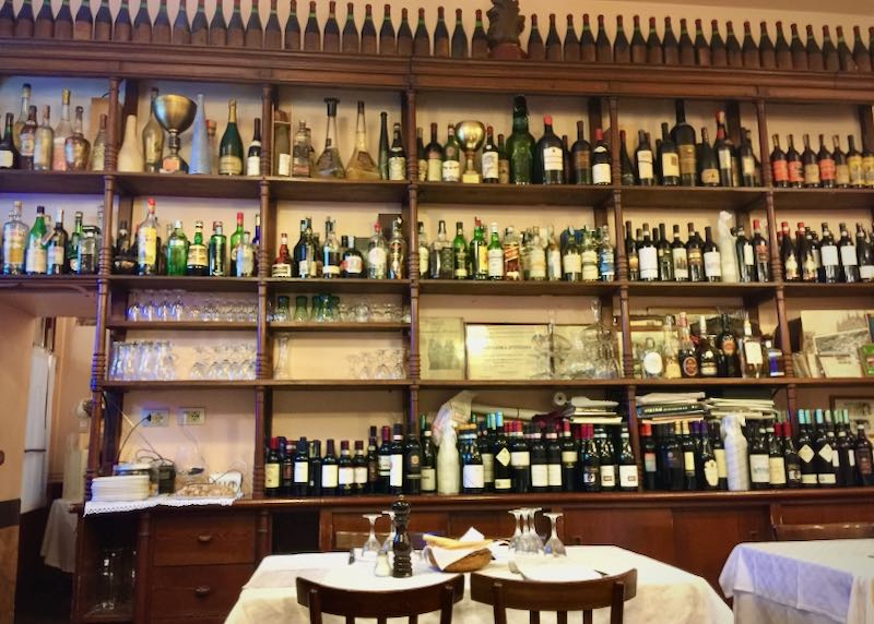 Dos sillas de madera y una mesa se colocan antes de los estantes de botellas en un restaurante italiano